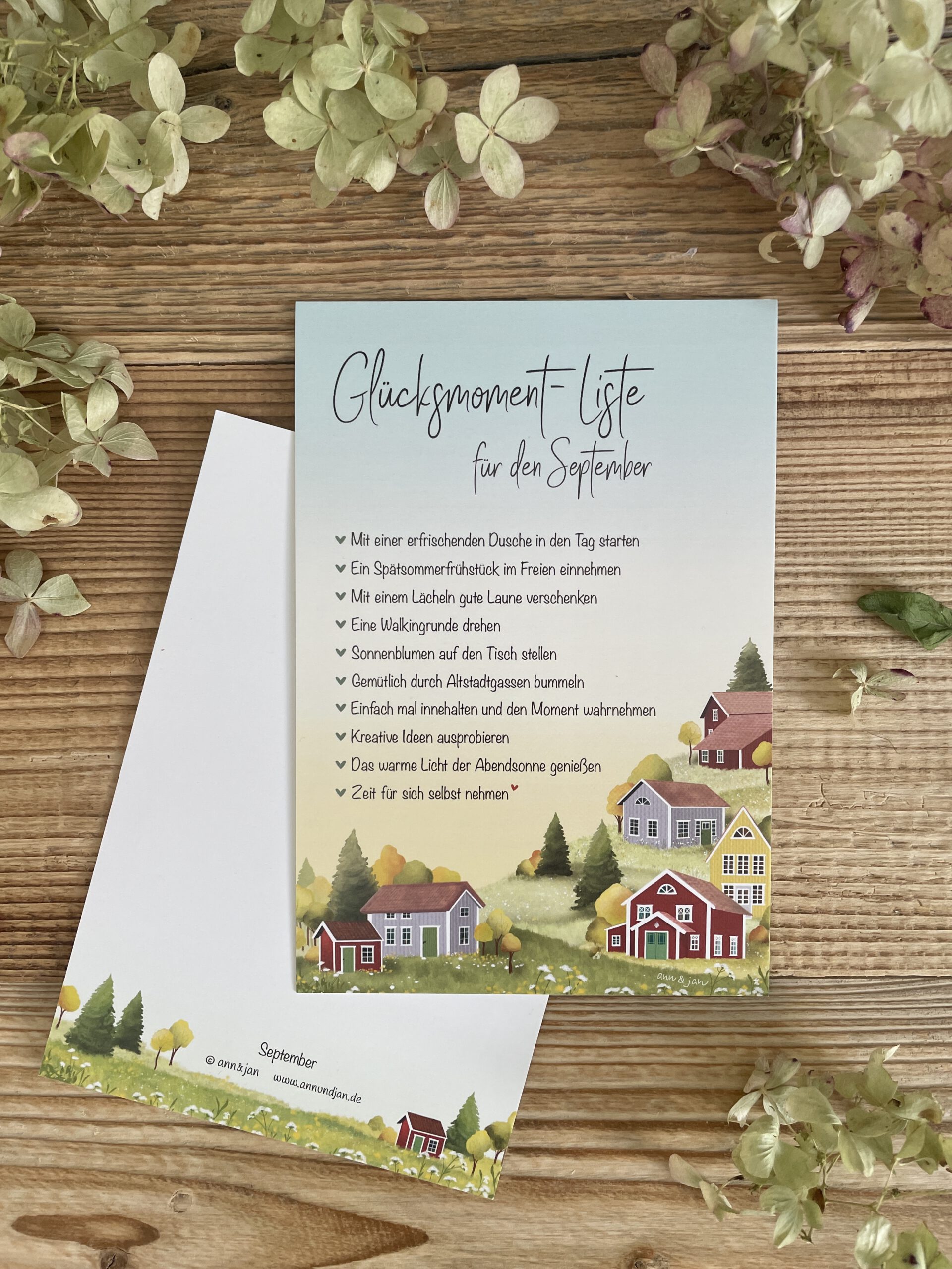 Handillustrierte große Postkarte Glücksmomente September bunte Holzhäuser Hügellandschaft buntes Laub Wiesenblumen von Ann und Jan